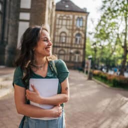 Smiling university student walking outside college campus holding notebook