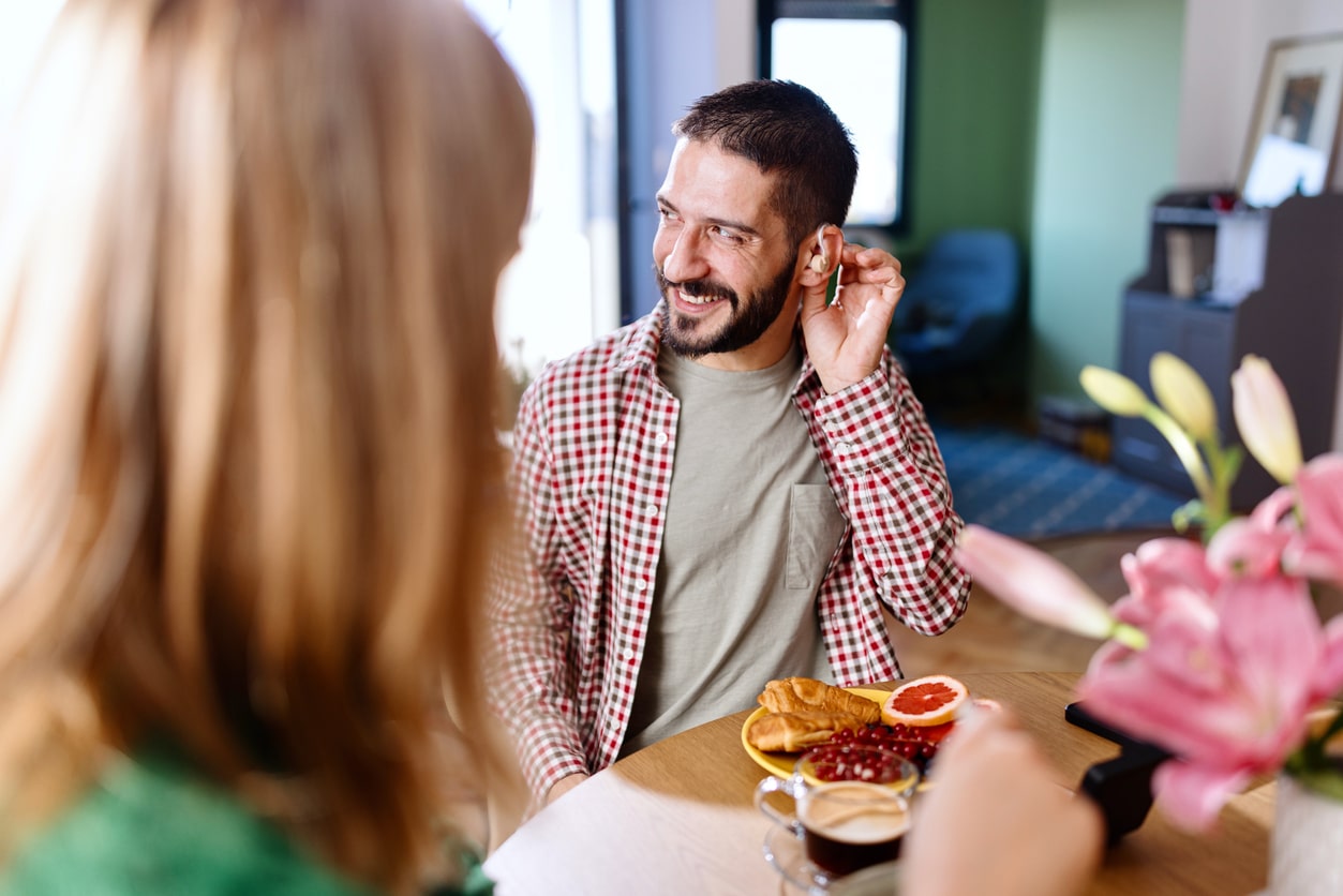Smiling man wearing audio aids while sitting at the kitchen table and talking with his family and friends.